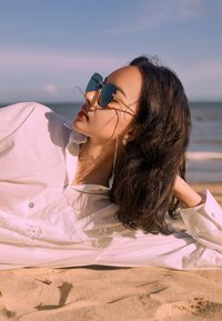 Eine Person, die im Sand liegt, trägt geometrische, blau getönte Sonnenbrillen, ein weißes, knopfbestücktes Hemd und einen silbernen Ohrring. Natürliche Kulisse des Ozeans.