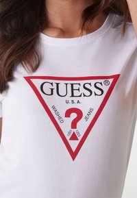 Camiseta blanca con un gran gráfico triangular rojo que incluye el texto "GUESS," "U.S.A." y "WASHED JEANS," además de un signo de interrogación rojo en el centro.