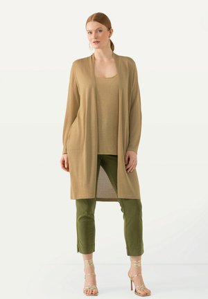 Beige dlouhý kardigan s kapsami přes béžový top, doplněný olivově zelenými úzkými kalhotami a sandály se zlatými pásky. Hladká textura.