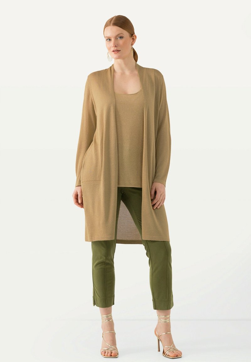 Cardigan long beige avec poches sur un top beige, assorti à un pantalon fitted vert olive et des sandales à brides dorées. Texture lisse.