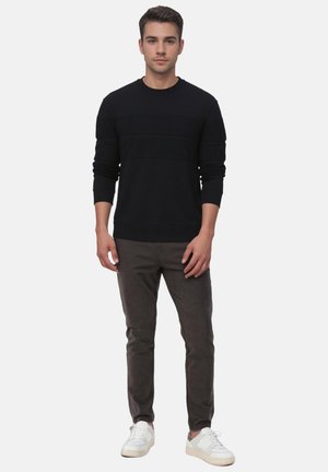 Junger Mann steht, trägt einen schwarzen Langarmpullover, dunkelgraue Hose und weiße Sneaker vor weißem Hintergrund.