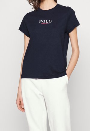 Kobieta ubrana w granatowy t-shirt z napisem "POLO Ralph Lauren" i białe spodnie, stoi z jedną ręką w kieszeni na jednolitym tle.