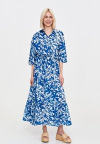 Floral maxi dress in blue and white with a collared neckline, cinched waist, tiered skirt, and three-quarter sleeves. Worn with platform sandals.