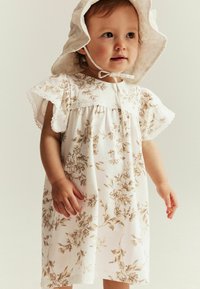 Weißes Kleid mit braunem Blumenmuster, kurzen Flatterärmeln, Spitzenverzierungen und einem passenden breitrandigen Hut, der unter dem Kinn gebunden ist.