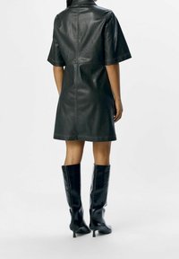 Robe en cuir noir avec des manches courtes, coupe au-dessus du genou et détails de couture. Assortie à des bottes noires à talons, montantes jusqu'aux genoux.