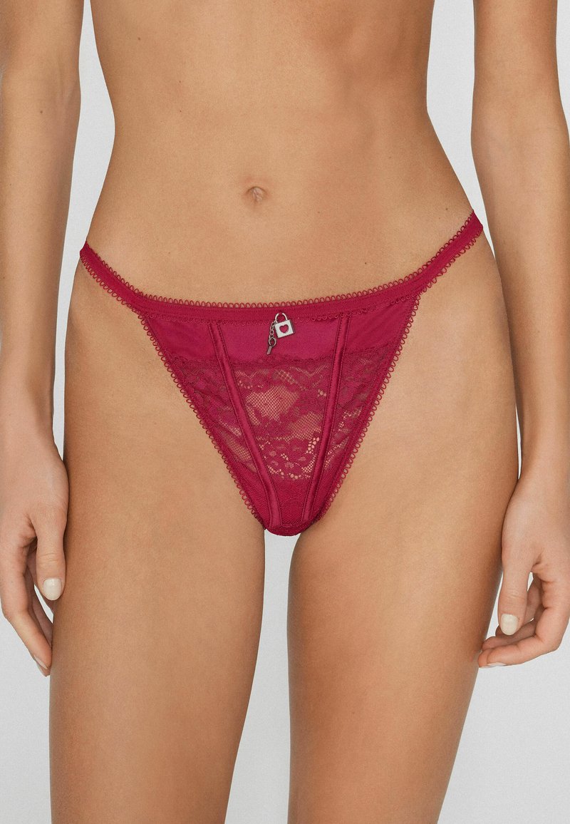 Kobieta w przezroczystych czerwonych stringach z koronki, z małym zawieszonym serduszkiem i kluczykiem na środku przedniego pasa.