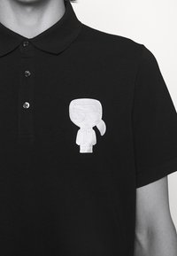 Černé polo tričko vyrobené z bavlny, s bílým vyšívaným designem postavy na levé straně hrudi. Detail se třemi knoflíky.