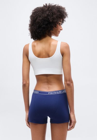 Γυναίκα με σγουρά μαλλιά που φοράει λευκό αθλητικό σουτιέν και μπλε σκούρα boxer Ralph Lauren Polo, στέκεται μπροστά σε ένα απλό φωτεινό φόντο.