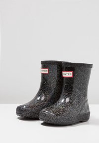 Botas de lluvia negras con brillo, hechas de goma. Cuentan con una punta redondeada, suela texturizada y una pequeña etiqueta con el logo rojo en la parte frontal.