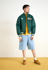Jachetă bomber din satin verde, cu manșete cu dungi albe și verzi, tricou galben, pantaloni scurți din denim bleu deschis și pantofi cu șireturi maro.