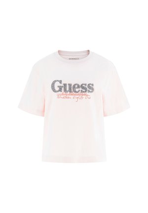 Roza pamučna majica s šljokicama koja prikazuje "Guess" logo, s natpisom "Established Nineteen Eighty One" u crvenoj boji, kratki rukavi i opušteni kroj.