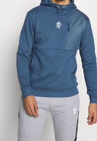 Felpa con cappuccio blu, dotata di una tasca frontale con zip, polsini elasticizzati e orlo a costine. Presenta un logo bianco sul petto e tasche laterali.