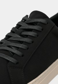 Svarte lær sneakers med flate svarte lisser, teksturert overflate og en kremfarget gummisåle. Fokuserer på minimalistisk design og glatte detaljer.