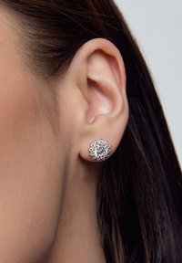 Srebrne kolczyki z wzorem kwiatu, ozdobione wieloma błyszczącymi, przejrzystymi kamieniami, ułożonymi w okrągły wzór, noszone w uchu.