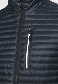 Marinblå isolerad jacka med horisontellt quiltat mönster, ståkrage, fullständig framdragkedja och en kontrasterande vit dragkedjeficka.