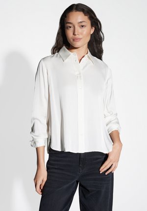 Camicia bianca a maniche lunghe, con bottoni, realizzata in tessuto morbido, con colletto a punta e vestibilità leggermente ampia, abbinata a jeans scuri.