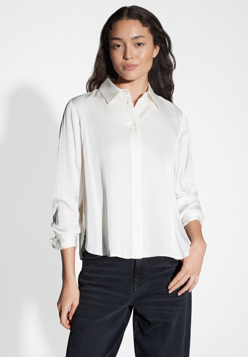 Camicia bianca a maniche lunghe, con bottoni, realizzata in tessuto morbido, con colletto a punta e vestibilità leggermente ampia, abbinata a jeans scuri.