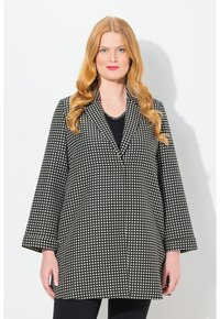 Manteau à pois noir et blanc avec col, manches larges et poches passepoilées, présentant une coupe droite et un tissu texturé.