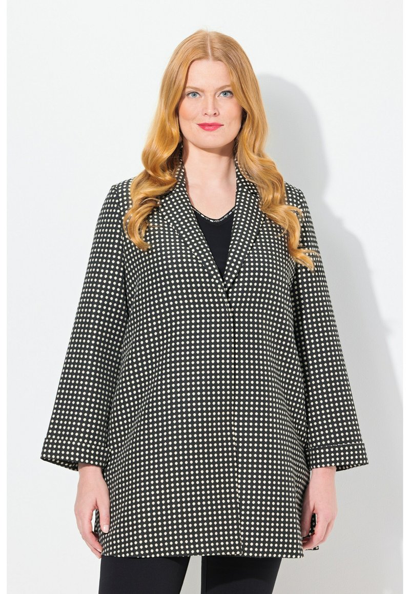 Manteau à pois noir et blanc avec col, manches larges et poches passepoilées, présentant une coupe droite et un tissu texturé.