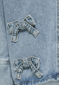 Světle modrá džínsová bunda se dvěma dekoračními textilními mašlemi přišity na přední straně. Denim má klasickou texturu a jemné detaily prošívání.