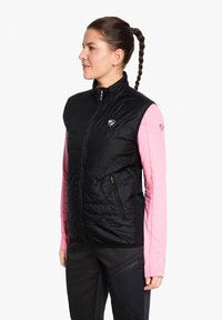 Black insulated vest with a high neck and zip closure, featuring diamond quilting, side pockets, and a logo on the chest. Worn over a pink long-sleeve top.