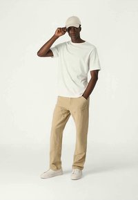 Homem a usar um chapéu branco, t-shirt branca, calças bege e ténis brancos, de pé com uma mão no bolso e a outra a ajustar o chapéu.