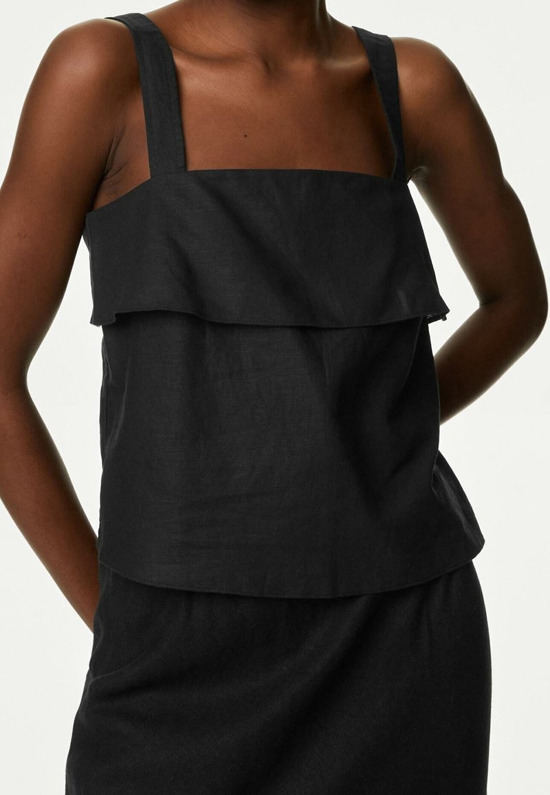 Femme portant un haut noir sans manches avec de larges bretelles et un détail volanté en couches sur la poitrine et un ourlet arrivant à la taille.
