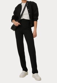 Person trägt eine schwarze Bomberjacke, ein weißes Hemd, schwarze hoch taillierte Hosen, weiße Sneakers und hält eine schwarze Umhängetasche.