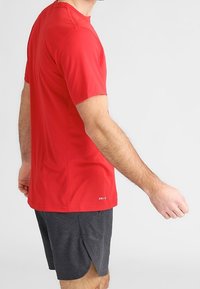 Hombre vistiendo una camiseta atlética de manga corta roja y pantalones cortos gris oscuro, de pie con el brazo extendido contra un fondo liso.