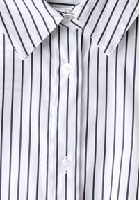 Valkoinen paita, jossa pystysuorat mustat raidat, terävä kaulus ja kaksi näkyvää kirkasta nappia napituslistassa.