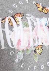 Grå bomull t-shirt med blommönster som visar rosa rosor och fjärilar i orange och svart. Vit text som stavar "HOLLISTER SOUTHERN CALIFORNIA."