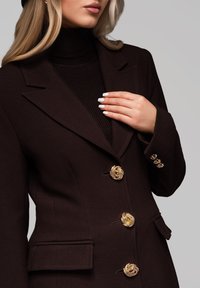 Brun skræddersyet blazer med gyldne udskårne knapper, klappede slag og sidelommer. Båret over en ribbet sort turtleneck.
