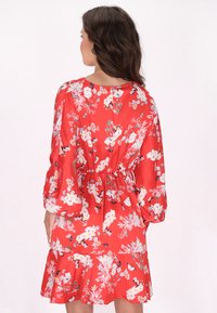 Rode bloemenjurk met korte ruches aan de onderkant, lange mouwen en elastische taille. Heeft verschillende lichtgekleurde bloemen- en vlinderpatronen.