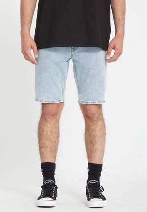 Hellblaue Jeansshorts mit geradem Schnitt, fransigem Saum und vier Taschen. Getragen mit schwarzen Sneakers und Socken in Knöchelhöhe.