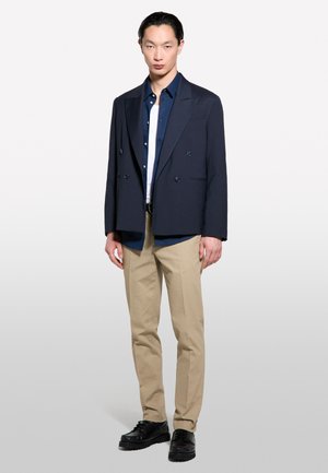 Blazer marine à double boutonnage sur une chemise marine à boutons et une chemise blanche, associé à un pantalon beige et des chaussures noires. Tissu lisse, coupe ajustée.