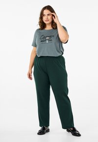 T-shirt gris avec un texte imprimé, associé à un pantalon ample de couleur vert foncé. Le modèle porte des mocassins noirs et a les cheveux jusqu'aux épaules.