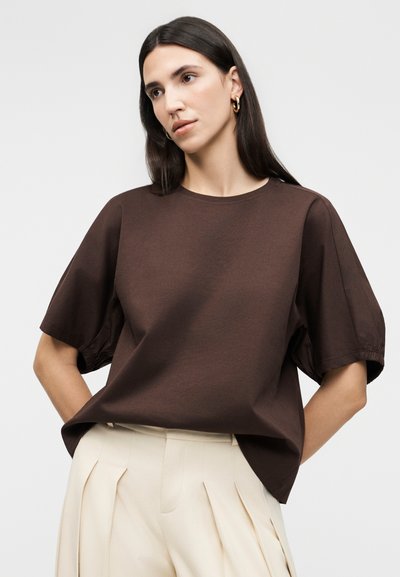 Γυναίκα με μακριά σκούρα μαλλιά που φορά χρυσούς κρίκους στα αυτιά, ένα φαρδύ καφέ κοντομάνικο τοπ και παντελόνι κρέμ με πιέτες και ψηλή μέση.