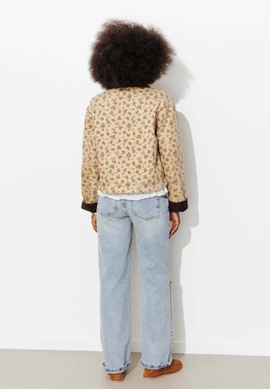 Personne aux cheveux bouclés se tenant face à un mur blanc, portant une veste beige à motifs floraux, un jean bleu clair et des chaussures marron.