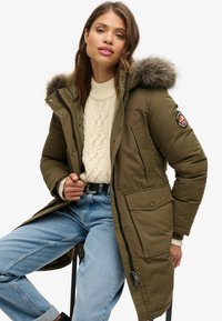 Parka verde oliva con capucha forrada de piel, cierre de cremallera y dos bolsillos frontales. Lleva puesta sobre un suéter de punto trenzado crema y unos vaqueros azules.