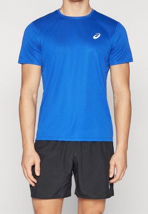 Man draagt een blauw sportshirt met korte mouwen en zwarte sportshorts, staat met ontspannen armen langs zijn zij.