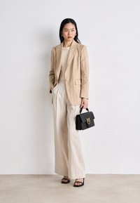 Béžový pruhovaný blazer cez krémový top, doplnený širokými ľanovými nohavicami. Nesie čiernu kabelku so zlatým kovanim a má na sebe čierne sandále.