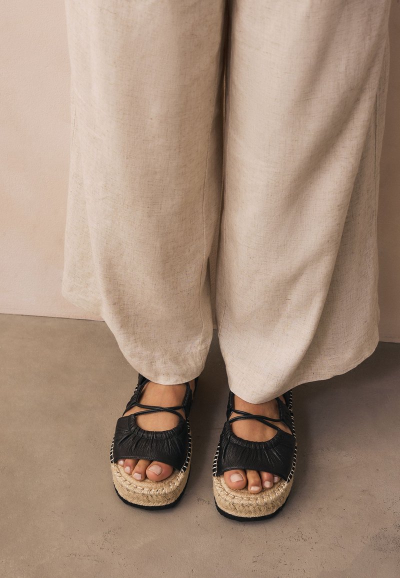 Sorte espadrillesandaler med tekstureret overdel og flettet jutesål, med et rundt design og krydsede stropper, båret med lysebeige bukser.