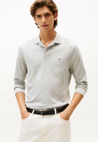 Polo a maniche lunghe grigio chiaro, realizzato in cotone, con colletto e un piccolo logo sul petto. Abbinato a pantaloni bianchi e una cintura intrecciata nera.