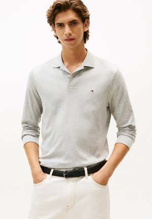 Polo shirt à manches longues gris clair, en coton, avec un col et un petit logo sur la poitrine. Assorti à un pantalon blanc et une ceinture tissée noire.
