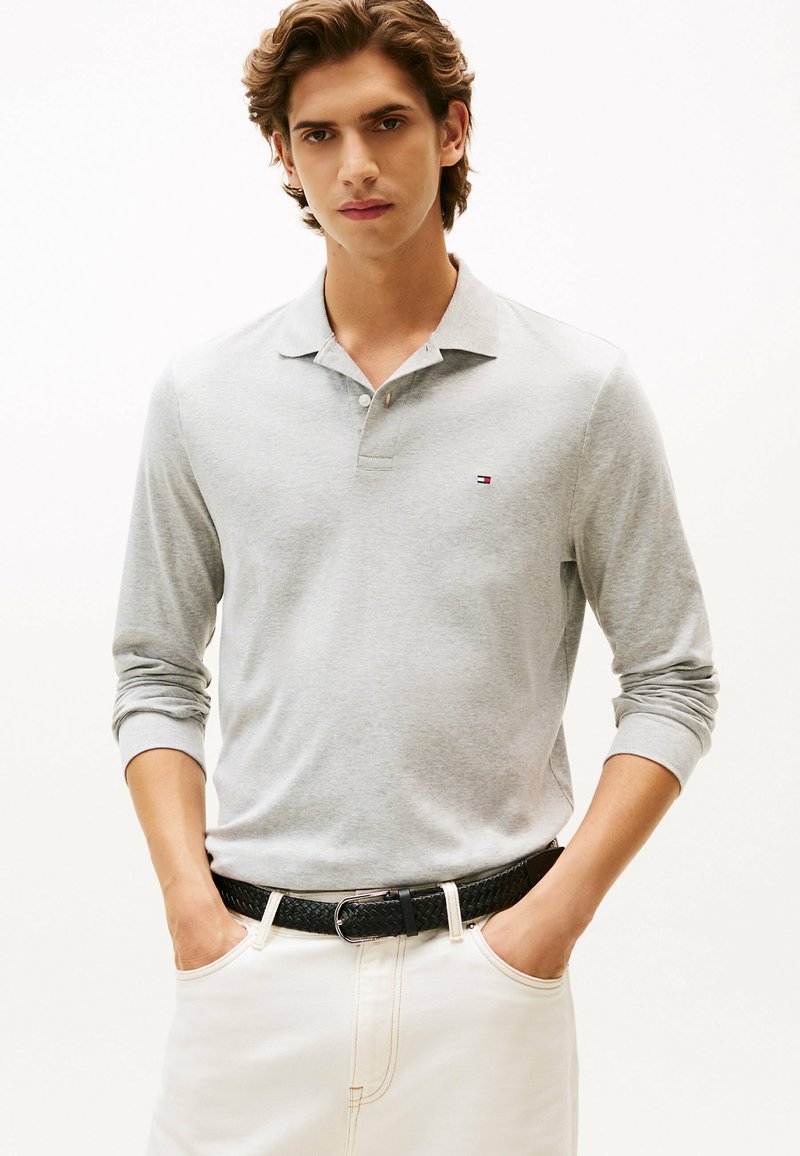 Polo a maniche lunghe grigio chiaro, realizzato in cotone, con colletto e un piccolo logo sul petto. Abbinato a pantaloni bianchi e una cintura intrecciata nera.