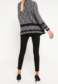 Svart och vit mönstrad kofta med långa ärmar, öppen framsida och en texturerad yta, kombinerad med svarta slim-fit byxor och högklackade skor.