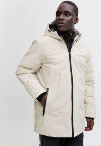 Mees, kes kannab beežikat kapuutsiga talvejopet musta luku ja taskutega, vaatab küljele tavalise tausta ees.