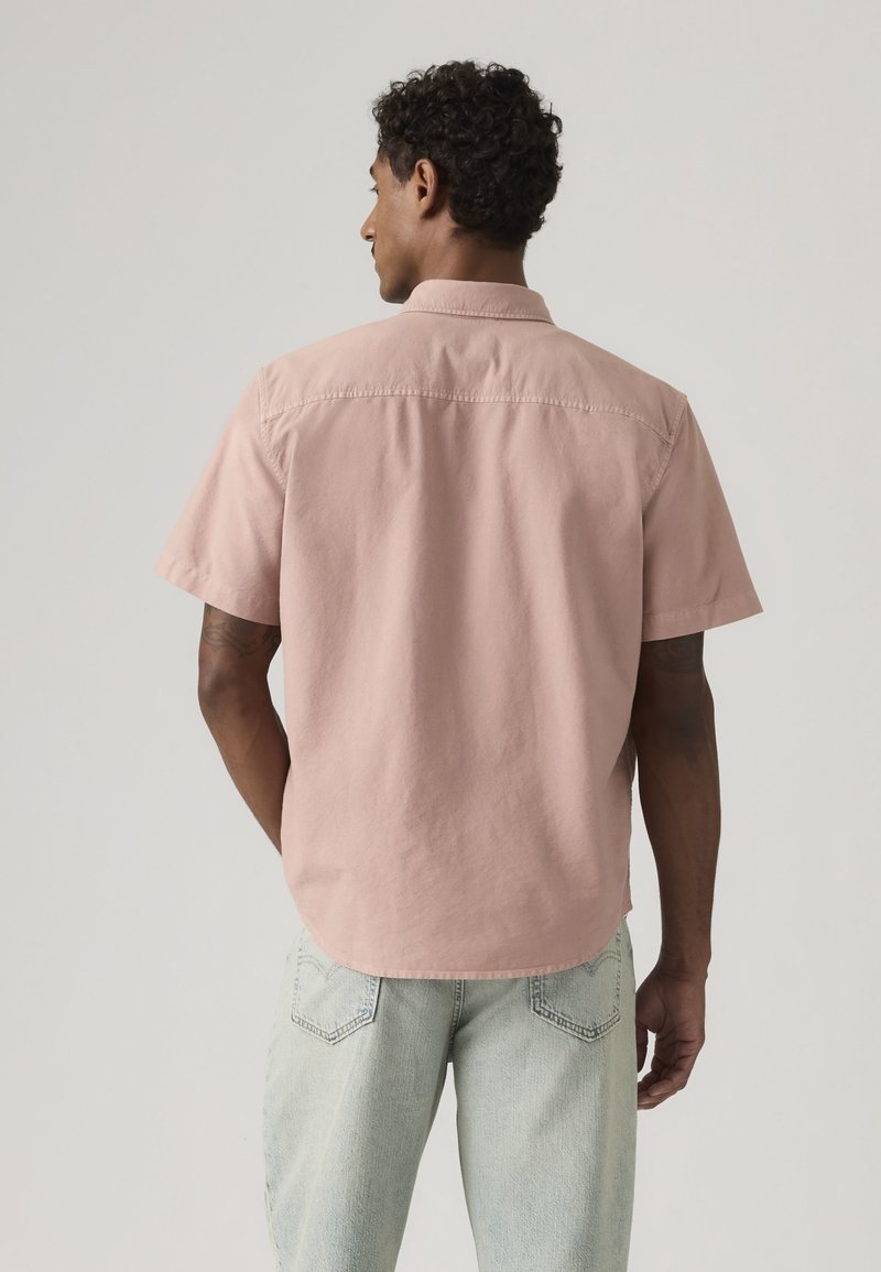 Mann mit lockigem Haar, von hinten gesehen, trägt ein hellrosa kurzärmeliges Hemd und hellblaue Jeans vor einem einfarbigen Hintergrund.