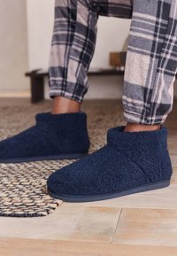 Pantofole blu, soffici e pelose, con design a metà caviglia e suola in gomma nera, abbinate a un abbigliamento da casa a quadri grigi e blu.