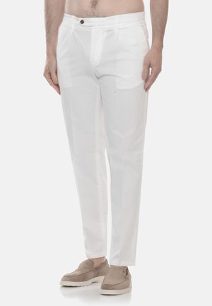 Uomo che indossa pantaloni bianchi slim fit e scarpe slip-on marrone chiaro, in piedi davanti a uno sfondo bianco uniforme.